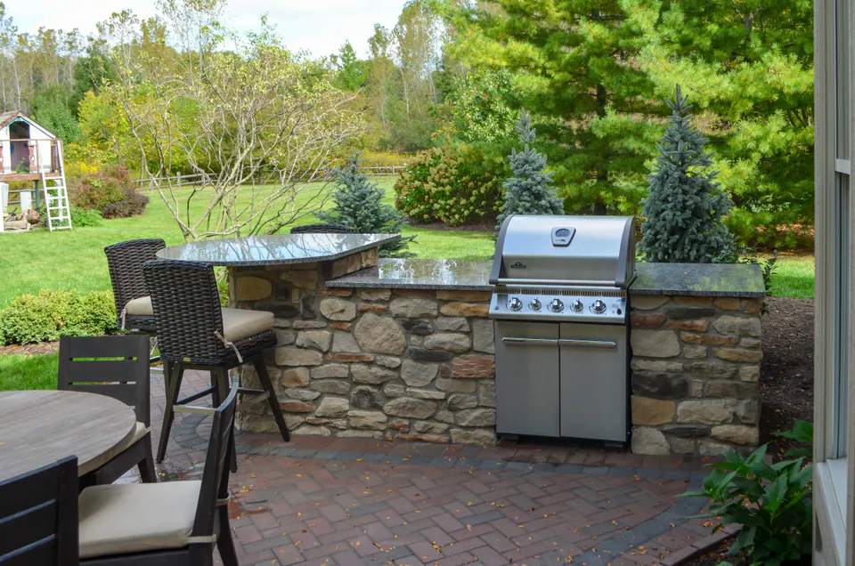 Outdoor grill station with stainless steel grill, natural stone wall base, paver patio, lounge seating, and wooded privacy planting